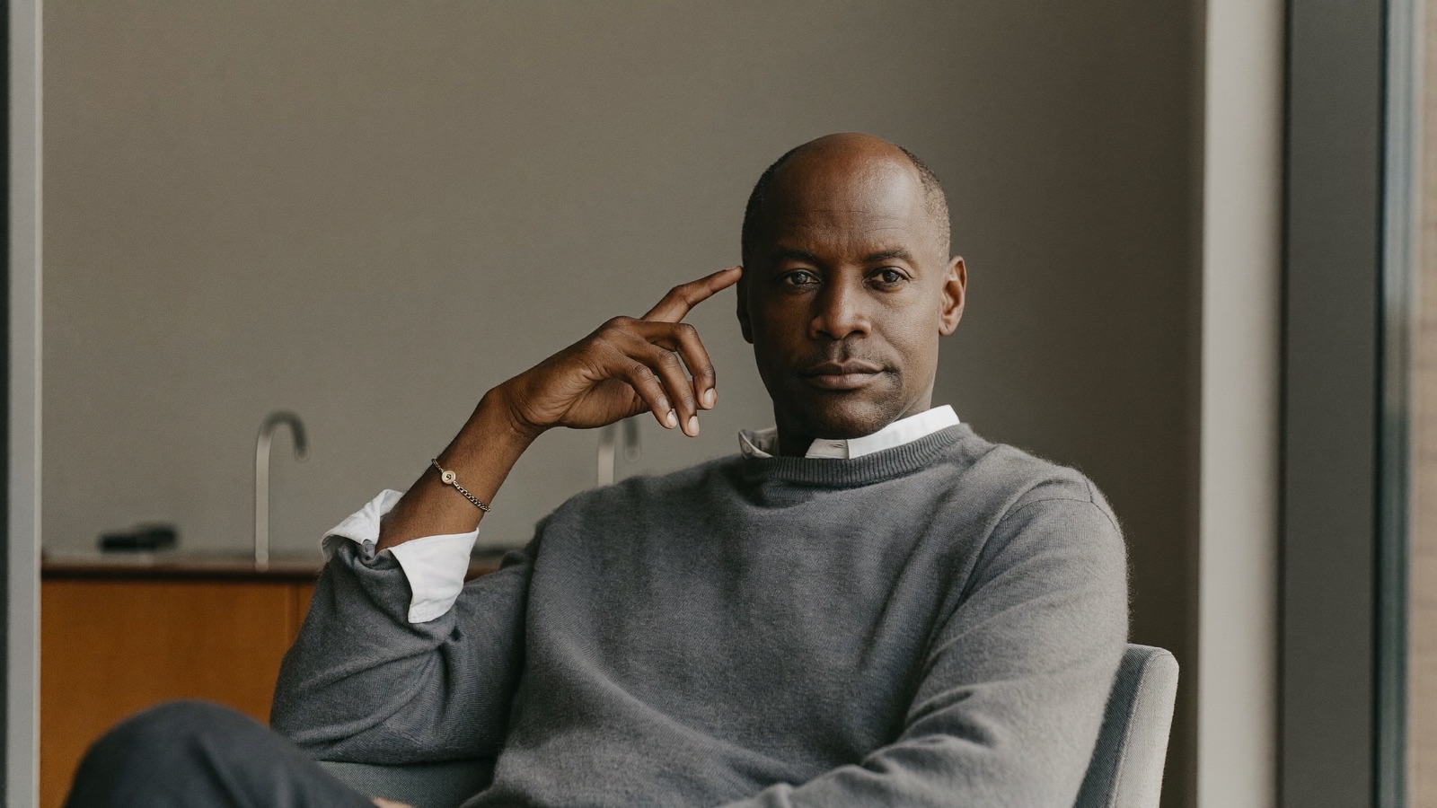Composed man in his fifties with advanced hair loss seated in a bright consultation room, resting one hand thoughtfully at his temple