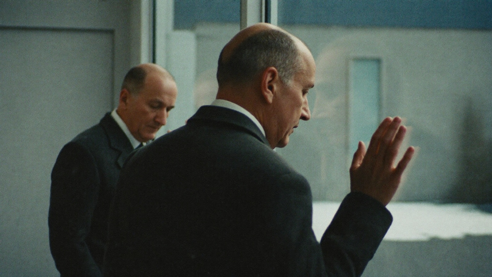 Man in a dark winter coat standing beside a window, his reflection visible in the glass, with snow outside