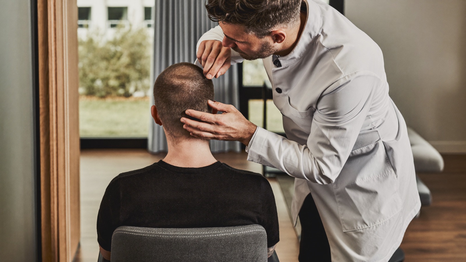 Clinician in a white coat examining the hair at the back of a seated patient's head during a consultation