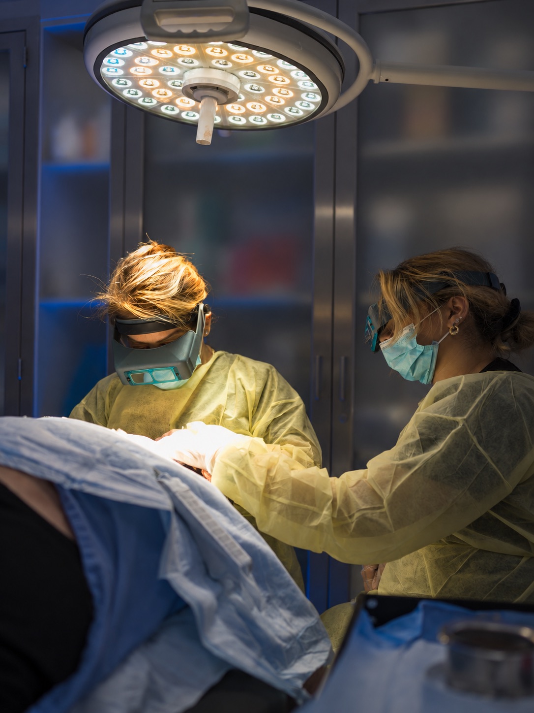 Two clinicians in protective gowns and magnifying loupes working under a surgical light during a transplant procedure