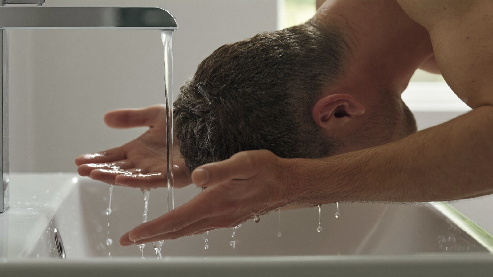 Man leaning over a bathroom sink holding his cupped hands beneath his short hair as water runs from the tap