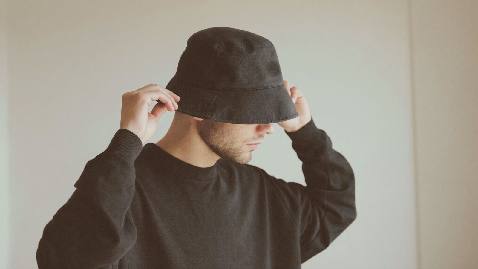 Man in a dark sweater gently lowering a plain loose cotton bucket hat onto his head with both hands against a bare white wall