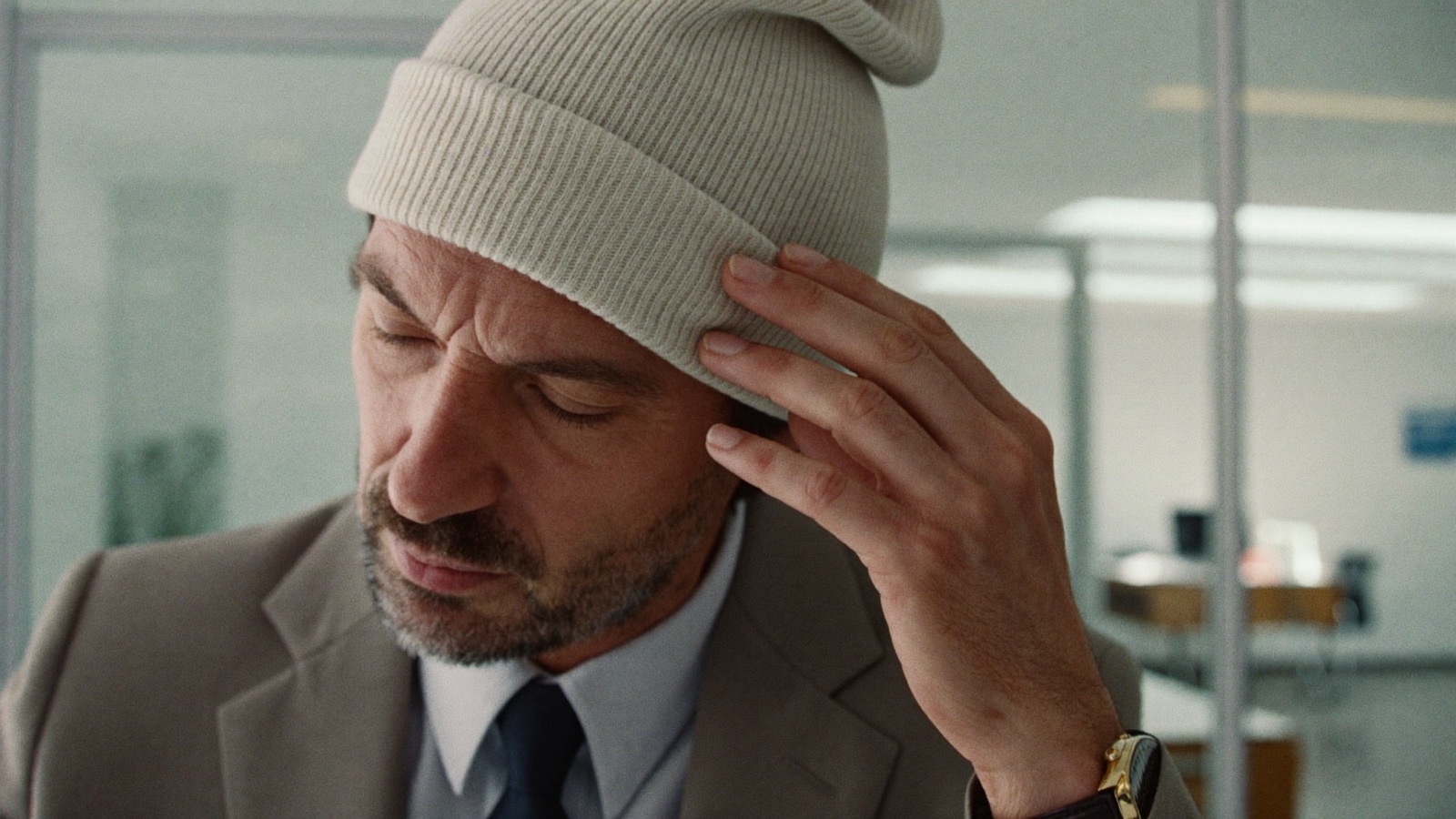 Man in a suit adjusting a cream beanie while standing in a glass-walled office