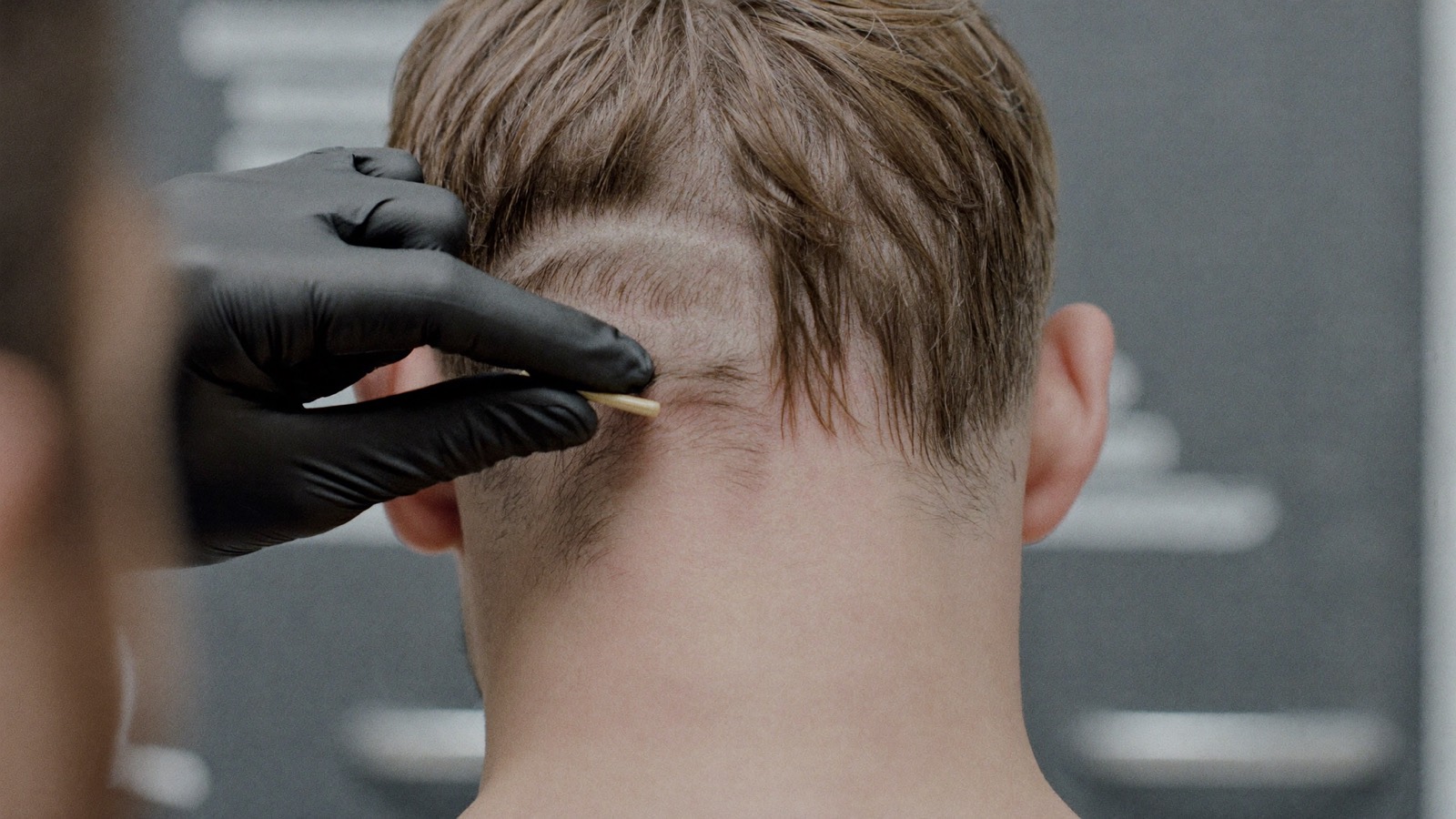Gloved hands lifting longer hair at the back of a man's head to reveal a narrow trimmed band beneath