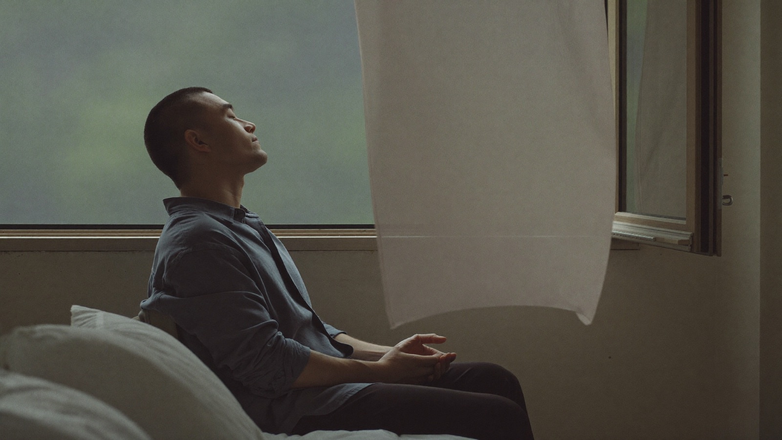 Man with a short buzzed haircut sitting calmly by an open bedroom window with his eyes closed and hands resting in his lap, cooling off during recovery after a hair transplant