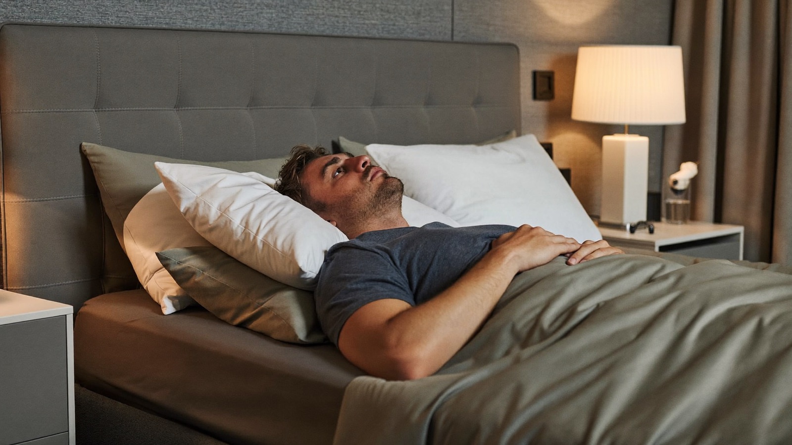 Man resting on his back propped up on stacked pillows in a lamp-lit bedroom at night, head raised and supported