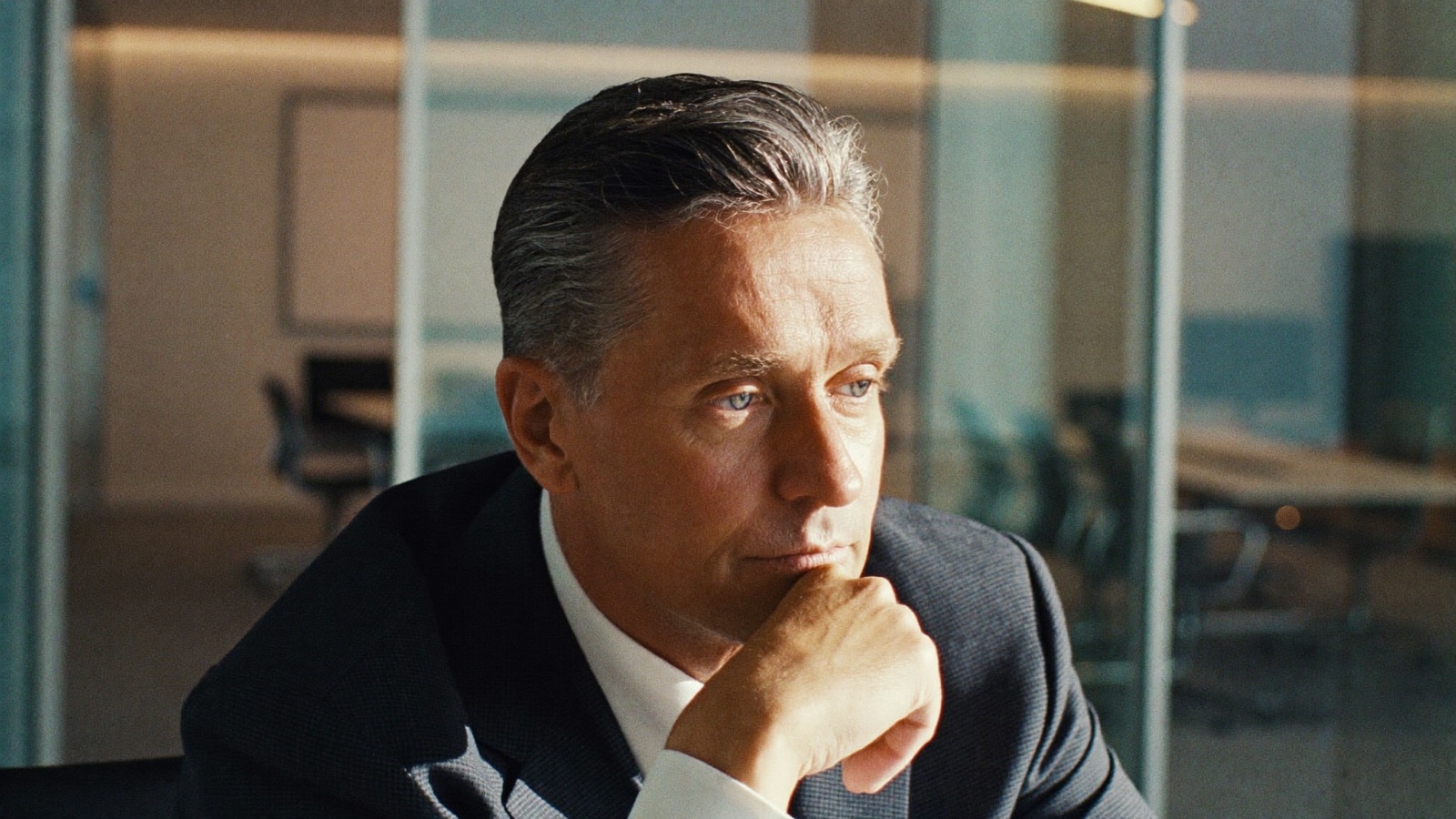 Man in a dark suit sitting thoughtfully in a glass-walled office meeting room